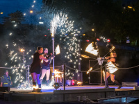 Drei Künstlerinnen führen nachts eine Feuershow mit Funken und brennenden Requisiten auf einer Bühne am Wasser auf.
